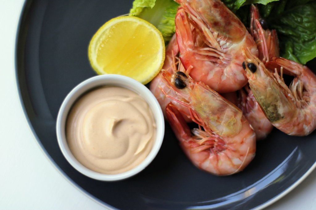 Keto prawn cocktail sauce in a white bowl on a black plate with unpeeled prawns and a lemon half next to it on the plate