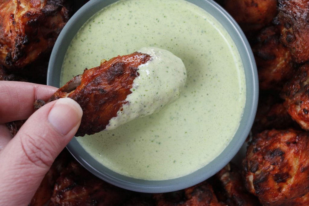 A hand holding a chicken wing dipped in the yoghurt sauce over the bowl of sauce.