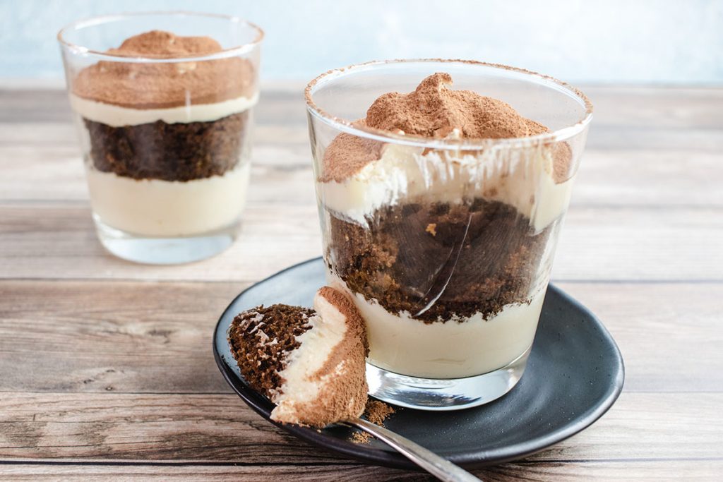 Keto tiramisu with a spoonful taken out on a black saucer. A silver spoon with some tiramisu on it rests on the plate. There is another tiramisu in the background. 