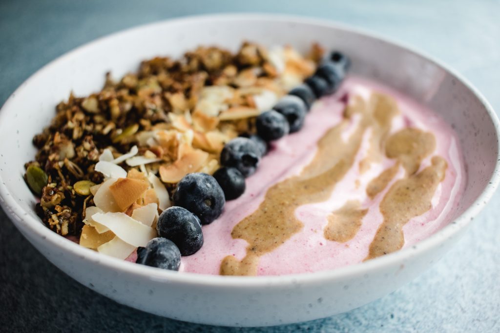 Strawberry smoothie bowl in a white speckled bowl topped with almond butter, blueberries, coconut flakes and granola. The toppings are all neatly in a line. 