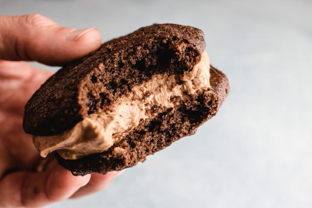 A hand holding a chocolate whoopie pie with a a bite out of it showing more of the filling. 