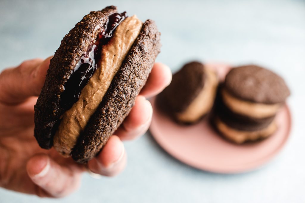 A hand holding one whoopie pie that is filled with jam with three whoopie pies on a pink plate in the background