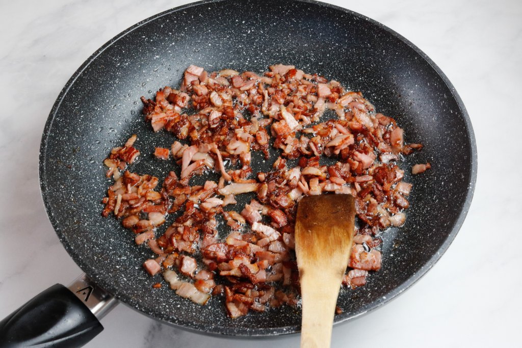 Diced bacon frying in a black speckled frying pan with a white background. 