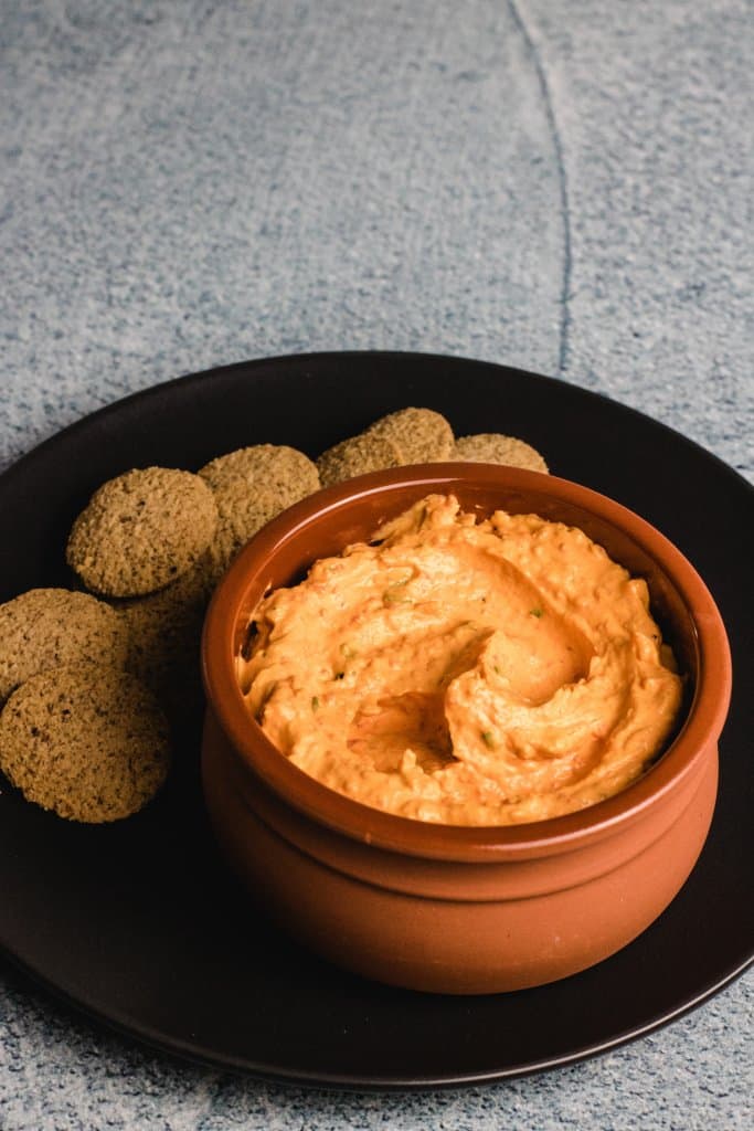 Roasted capsicum dip in a terracotta bowl sitting on a black plate with round crackers beside the dip.