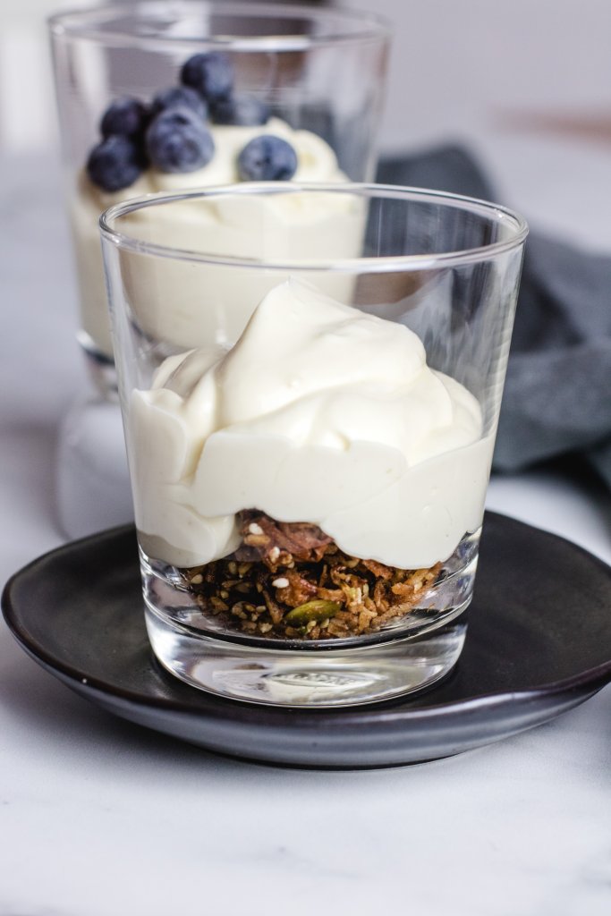 Keto cheesecake fluff on top of some granola  in a clear glass, sitting on a small black plate with another cheesecake fluff in a glass behind it. 