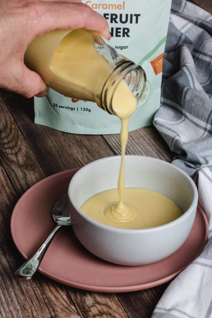 keto salted caramel custard in a white bowl, sitting on a pink plate with a silver spoon sitting on the plate. A small bottle of custard is being poured into the bowl. A locako packet and a grey checked tea towel in the background. 