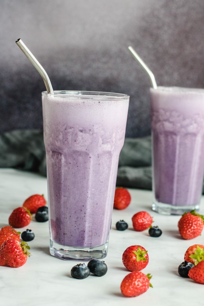 two glasses with breakfast smoothie in them and metal straws with strawberries and blueberries scattered around. 