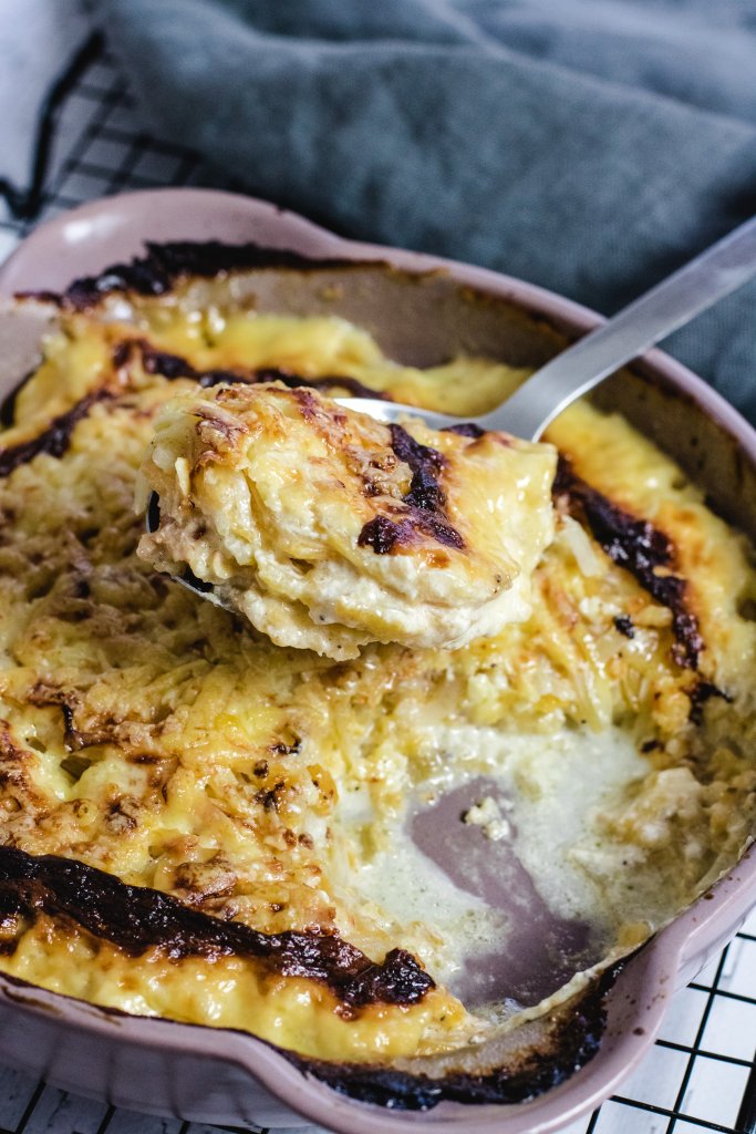 Keto potato bake in a pink dish on a wire rack on a white wooden background with a metal spoon lifting some of the potato bake on it. 
