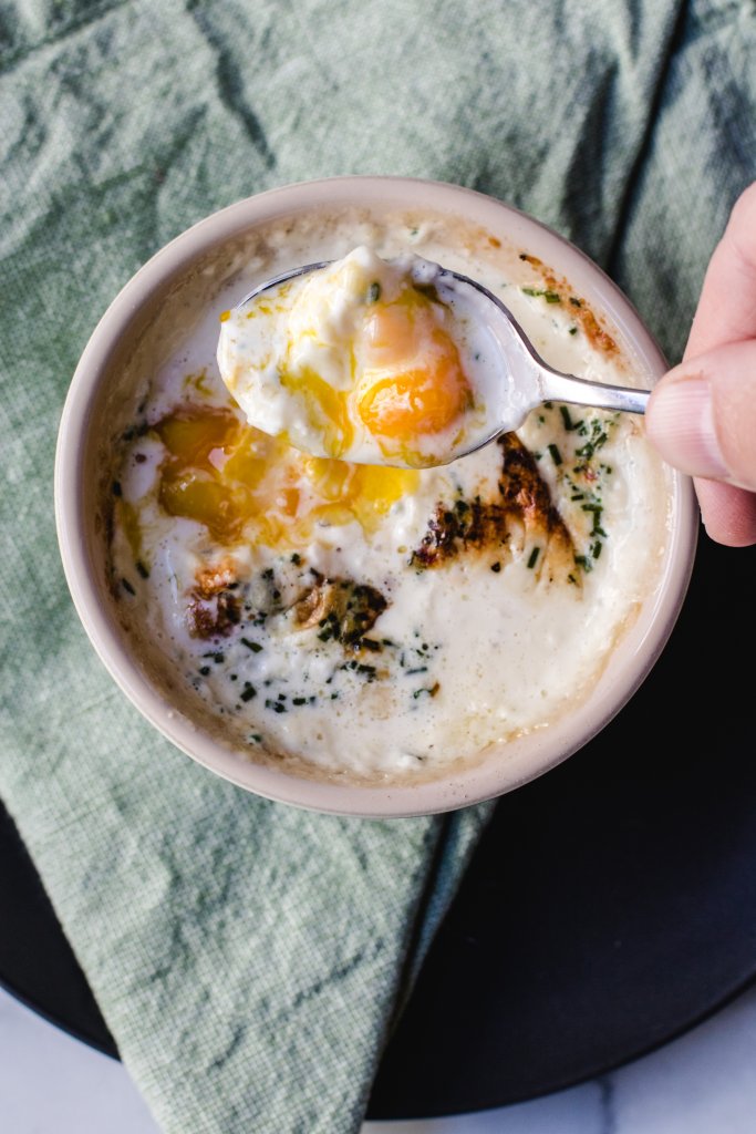 A ramekin full of creamy baked eggs sitting on a napkin. There is a hand with a spoon scooping out the runny yolk.