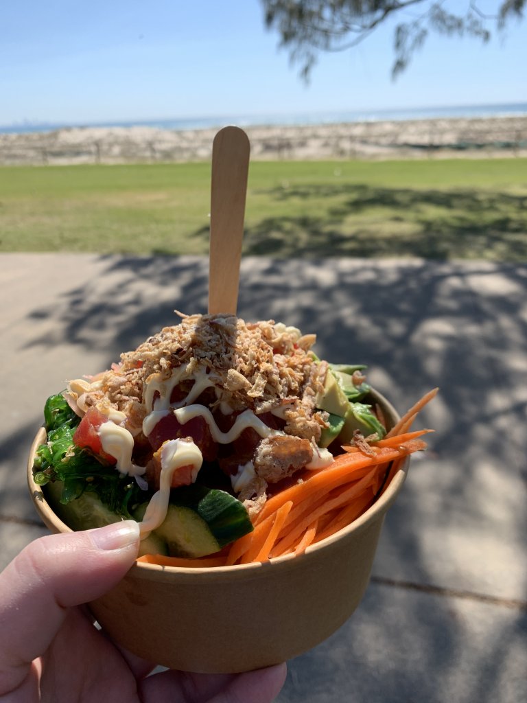 A poke salad bowl in a takeaway bowl with a bamboo fork. The bowl is held in one hand with some grass and then the beach and ocean in the background.