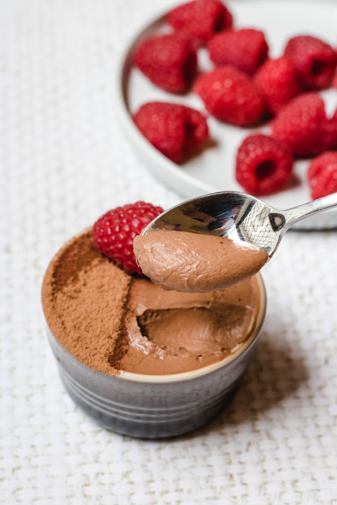 Keto chocolate mousse in a gray ramekin, topped with a raspberry. A spoon above with a small amount mousse on it. 