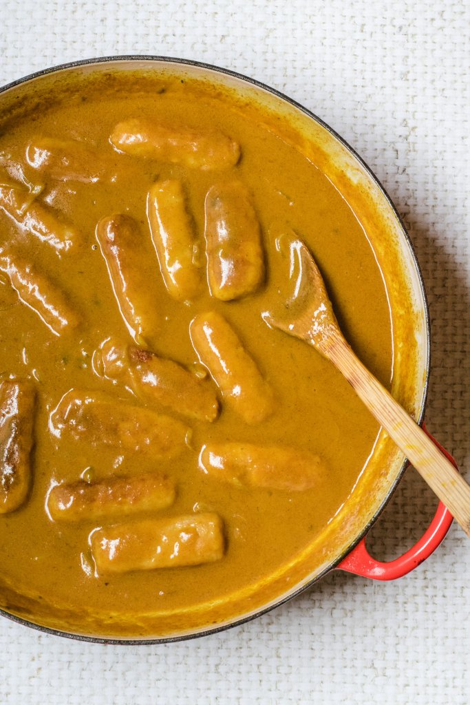 Keto curried sausages in a red pan ith a wooden spoon in it