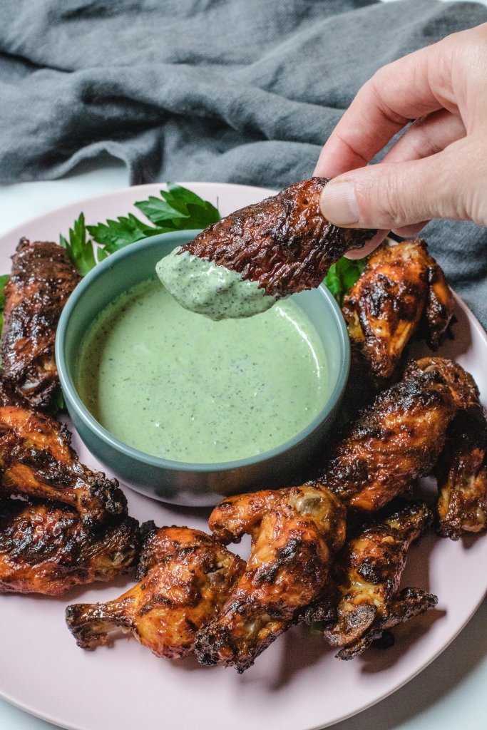 A hand holding a chicken wing dipped in the yoghurt sauce over the bowl of sauce.