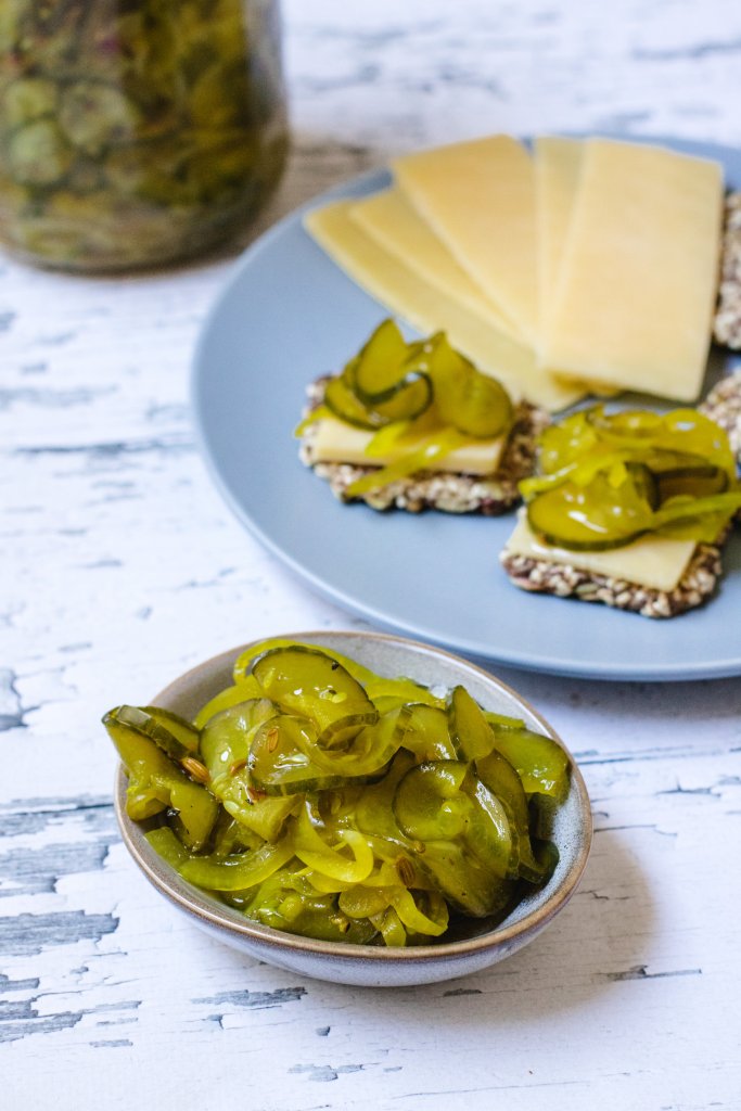 A small bowl of pickles on a white wood background with a large jar of pickles in the background and a plate with crackers, cheese and pickles. 