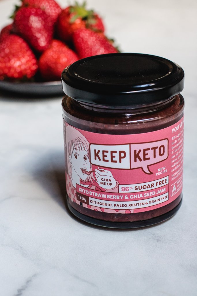 A jar of keep keto keto strawberry and chia seed jam with strawberries on a black plate in the background