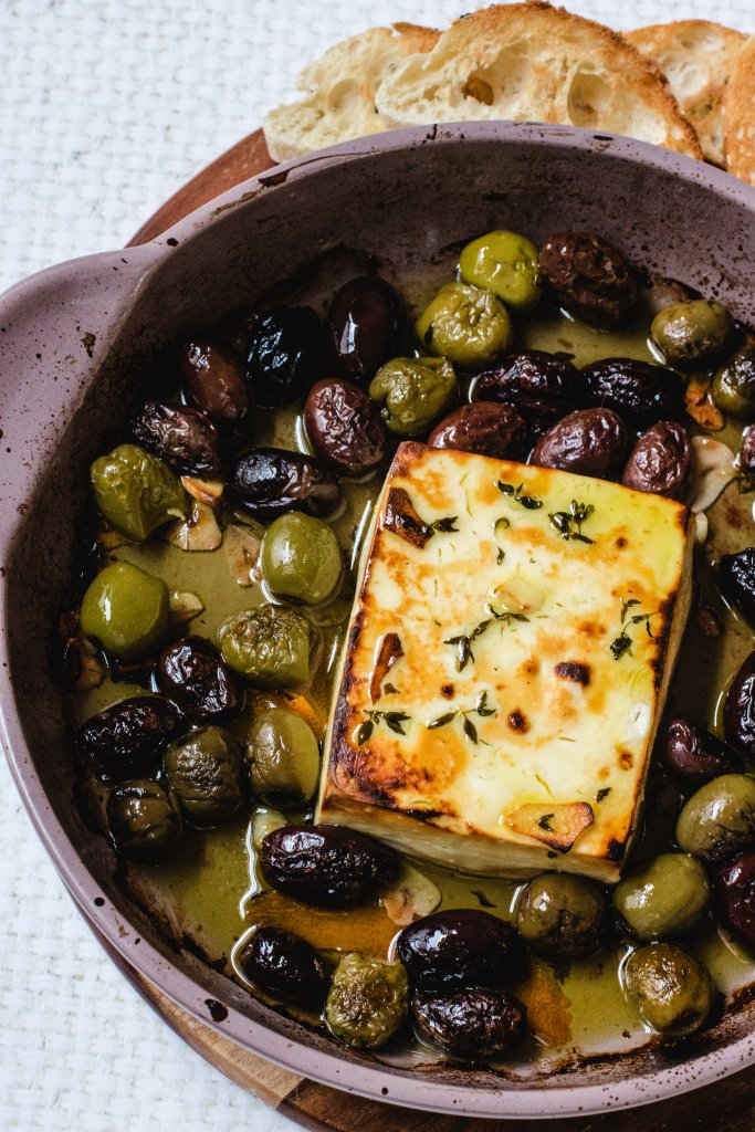 An overhead shot of baked feta and olives in a baking dish with toasted Turkish bread pieces around the edge.
