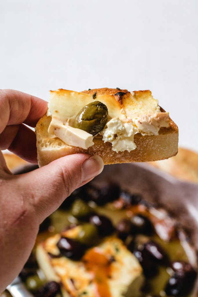 A hand holding up a piece of toasted Turkish bread spread with the baked feta and olives.