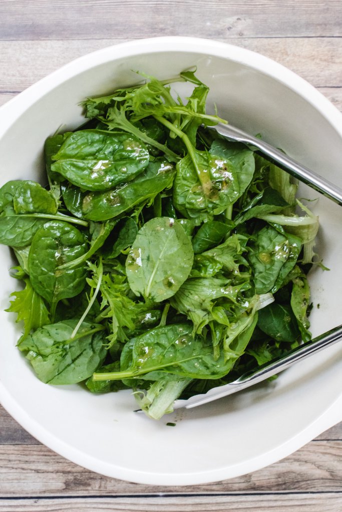 Mixed salad leaves with dressing in a white bowl with silver tongs on a white background