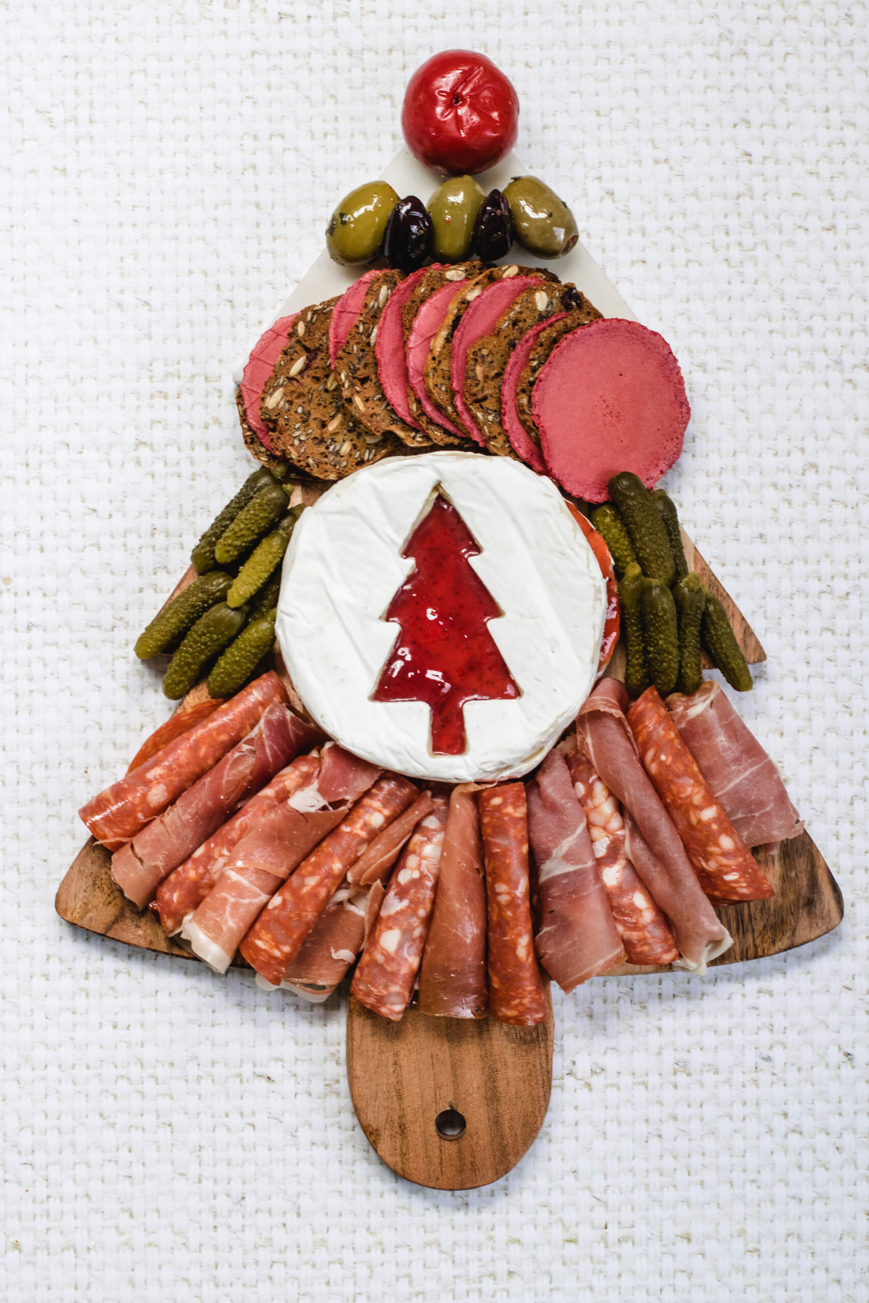 Christmas charcuterie board in the shape of a Christmas tree with beetroot crackers, brie with a christmas tree cut into it and deli meats, gerkins and olives. 