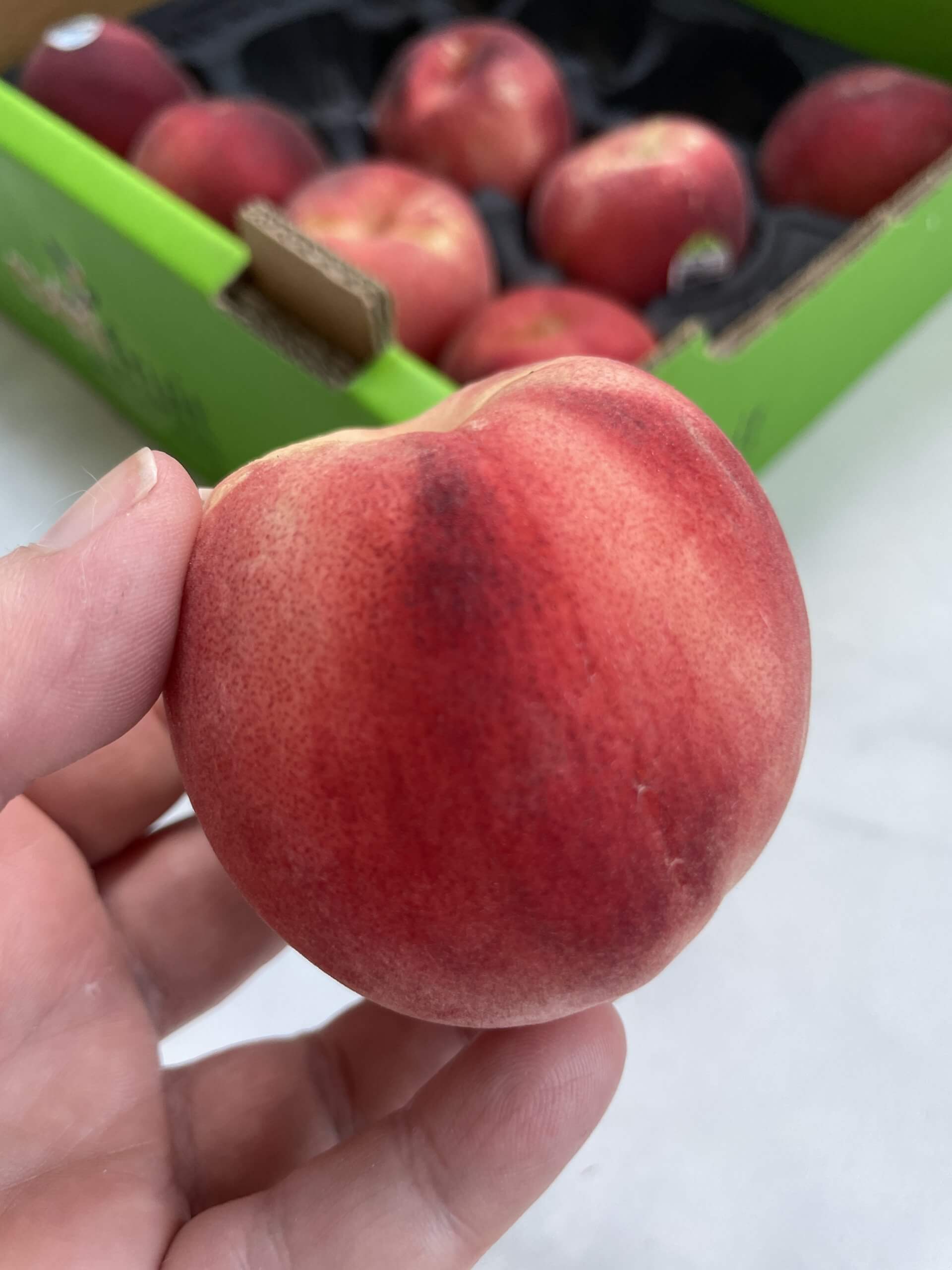 A hand holding a white peach with a box of peaches in the background. 